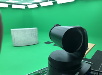 Blick von der Regie des GreenScreenStudios in den Aufnahmeraum. Im Vordergrund sieht man die PTZ-Kamera. Im Aufnahmeraum, dessen Wände grün gestrichen sind, steht ein Tresen in Betonoptik.