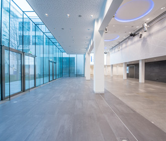 Lichtdurchflutetes Foyer mit deckenhohen Glasscheiben im Erdgeschoß im Haus der Bayerischen Wirtschaft. Das Bild zeigt den großen Eingangsbereich mit weißen Säulen und blauer Ringbeleuchtung